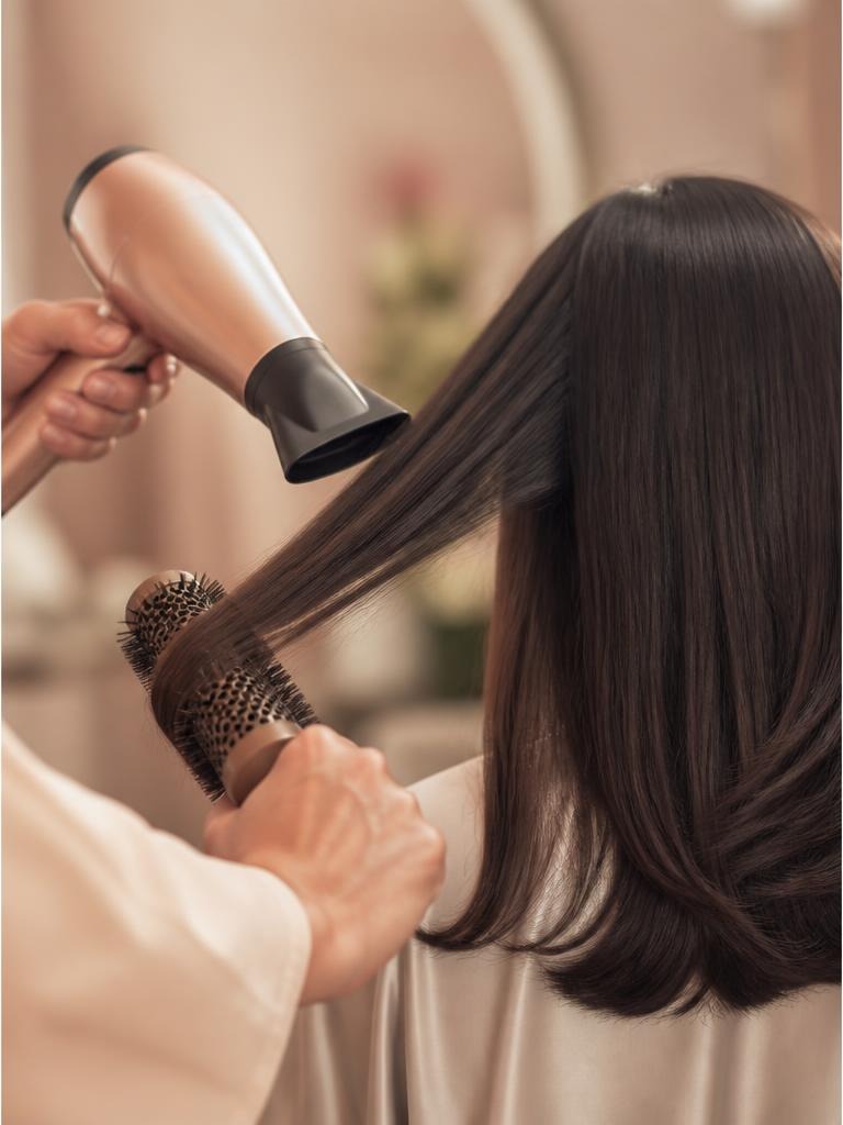 Stylist blow-drying long dark hair with a round brush during a salon appointment.
