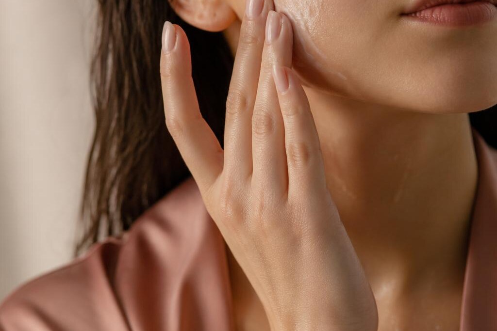 Person gently pressing moisturiser into a cheek in warm, soft light.